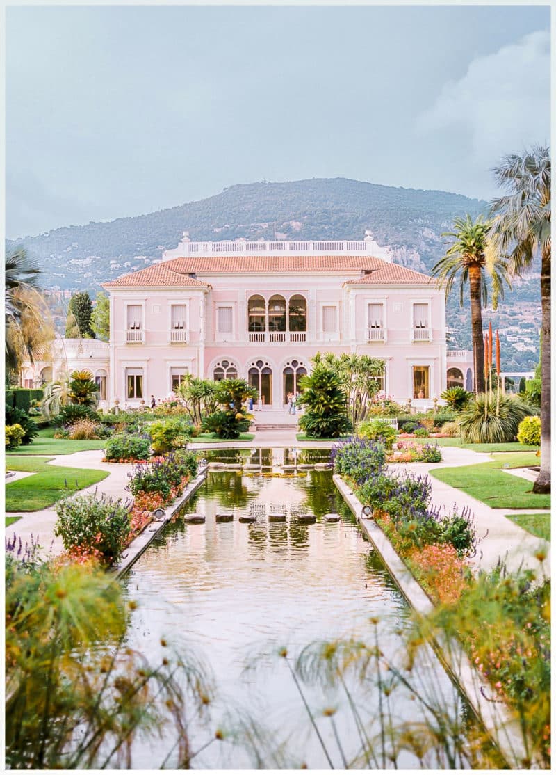Villa Ephrussi de Rothschild rose-coloured Belle Époque façade reflected in the Florentine garden's ornamental pool, with palm trees and the French Riviera hills behind — Saint-Jean-Cap-Ferrat, France