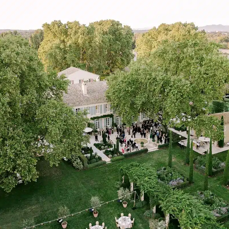 Aerial drone view of a wedding celebration at a Provence château estate — guests gathered in the formal gardens with the limestone manor house surrounded by mature trees — HD Moments