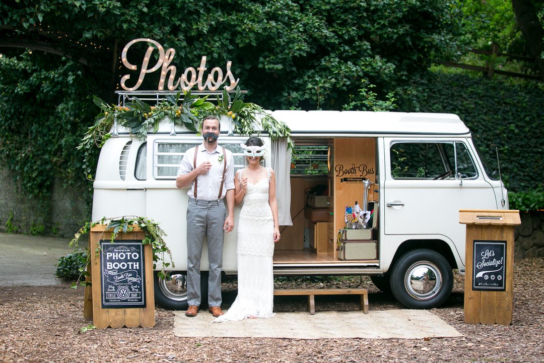 Bride and groom standing in front of a vintage VW campervan photo booth decorated with a neon Photos sign and fresh florals — unique wedding entertainment idea 2026 — HD Moments