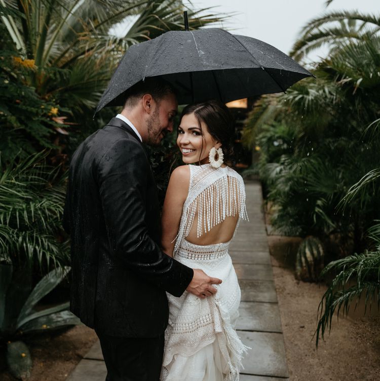 Bride smiling over her shoulder in a stunning open-back pearl fringe wedding gown, sheltering under an umbrella with her groom among lush tropical palms on their rainy wedding day — HD Moments