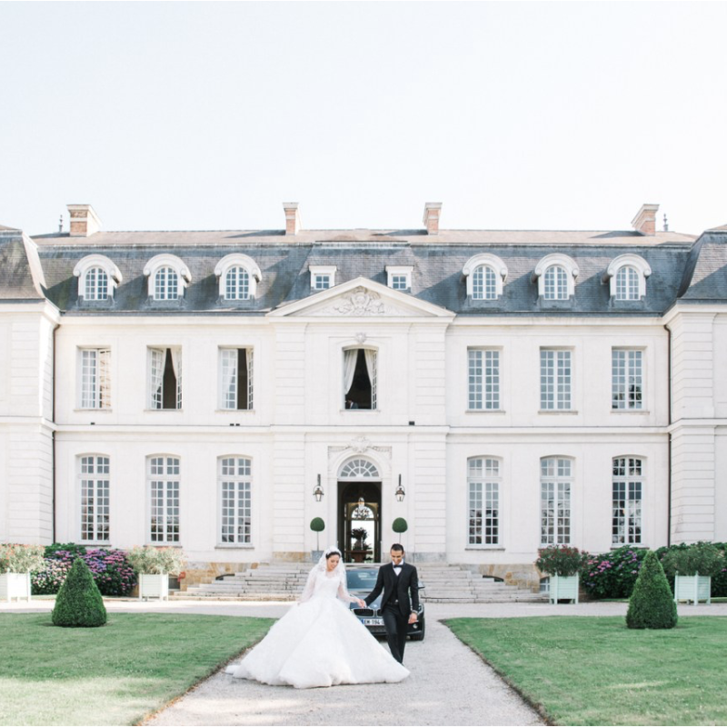 Bride Jumana in a voluminous white lace wedding gown and groom Ryan in a black tuxedo walking together in front of the neoclassical façade of Château du Grand-Lucé, Loire Valley, France — HD Moments destination wedding videographers